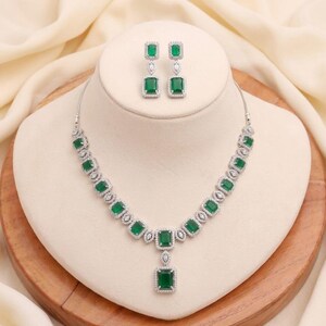 May include: A silver-toned necklace and matching earrings set. The jewelry features emerald-green square-cut stones surrounded by smaller clear stones. The necklace is displayed on a white bust, set against a cream-colored fabric.