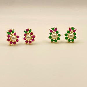 Może przedstawiać: Złote kolczyki sztyfty o kwiatowym wzorze. Każdy kolczyk ma centralną perłę otoczoną naprzemiennie czerwonymi i zielonymi kamieniami szlachetnymi. Kolczyki są umieszczone na neutralnym tle.
