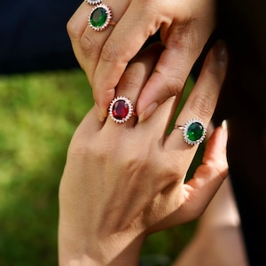 May include: Four rings with oval-shaped gemstones. Two rings have red gemstones, one ring has a green gemstone, and one ring has a dark green gemstone. All rings have a halo of smaller clear gemstones.