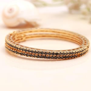 Puede incluir: Una pulsera tipo brazalete de tono dorado con dos filas paralelas de pequeñas piedras negras facetadas incrustadas en la banda metálica. La pieza de joyería circular presenta un diseño estructurado y calado en los laterales.