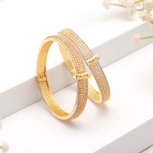 May include: Two gold-colored bangles with a textured, dotted pattern. The bangles have a clasp closure and are set against a white background. The jewelry is a classic accessory.