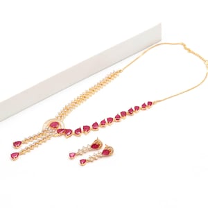 Puede incluir: Un conjunto de collar y pendientes en tono dorado con piedras rojas en forma de lágrima y cristales blancos transparentes. El collar presenta un motivo circular central con una piedra roja, extendiéndose en dos hileras de cristales y piedras.