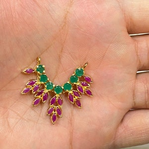 May include: A gold-toned metal jewelry connector featuring a fan-shaped arrangement of round green stones and marquise-cut fuchsia stones. The piece has two small loops at the top for attachment and a symmetrical design with a polished metallic finish.