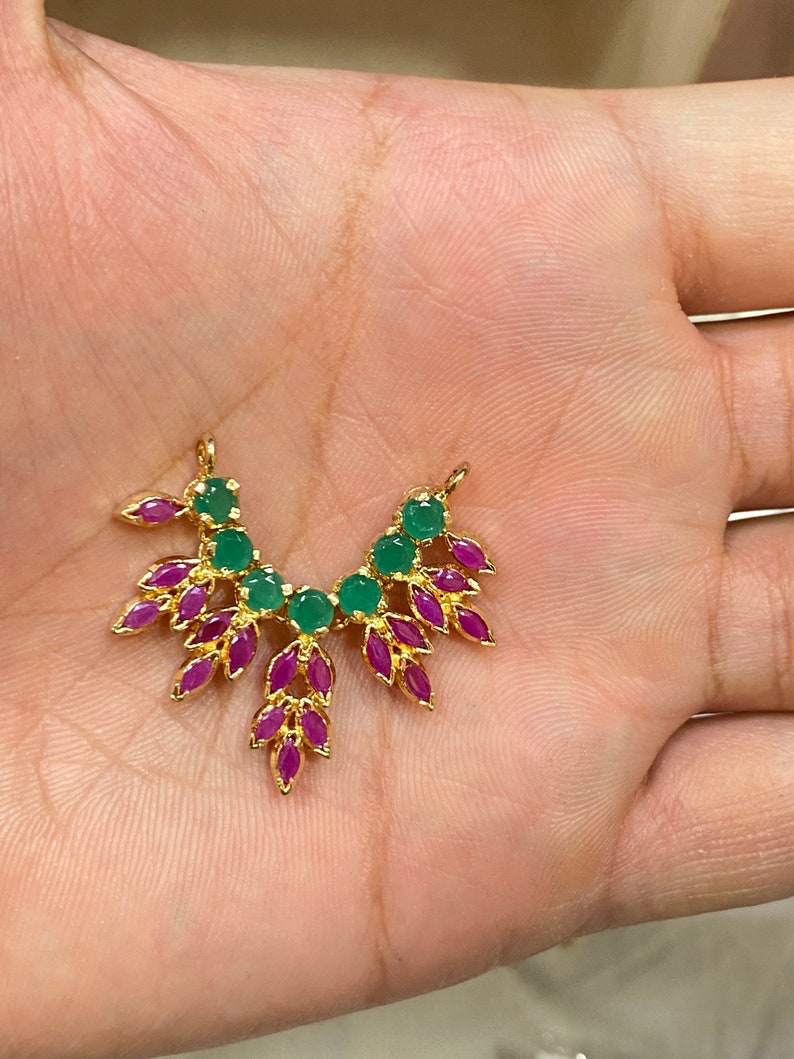 May include: A gold-toned metal jewelry connector featuring a fan-shaped arrangement of round green stones and marquise-cut fuchsia stones. The piece has two small loops at the top for attachment and a symmetrical design with a polished metallic finish.