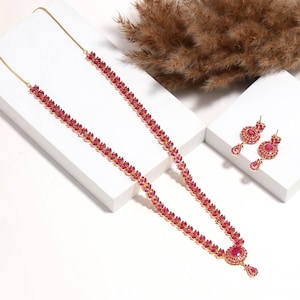 Puede incluir: Un conjunto de collar y pendientes a juego en tono dorado. El collar presenta una serie de piedras preciosas rojas en un patrón similar a una hoja, que culmina en un colgante más grande. Los pendientes tienen un diseño similar, con una parte superior redonda.