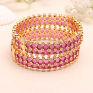 Puede incluir: Una pulsera dorada con un diseño de piedras preciosas rosas. La pulsera presenta filas de piedras preciosas rosas y pequeñas piedras transparentes. La pulsera es de estilo bisagra y parece ser de metal.