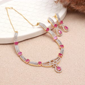 Peut inclure: Un ensemble collier et boucles d'oreilles doré orné de multiples pierres roses ovales et de cristaux pavés transparents. Le collier présente un design à plusieurs rangs avec un pendentif central en goutte, et les boucles sont pendantes.