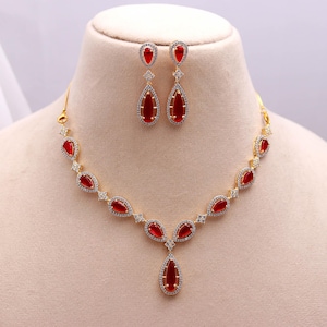 May include: A gold-toned necklace and earring set featuring teardrop-shaped red gemstones. The necklace and earrings are adorned with small, clear accent stones. The jewelry is displayed on a mannequin.