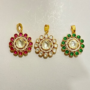 May include: Three gold pendants with a round design. The pendants have a central white stone surrounded by smaller white stones. The outer ring of stones is pink, white, and green.