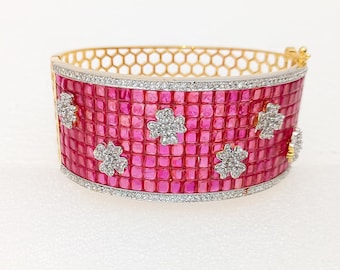 May include: A wide gold-tone bracelet with a pink, square-cut stone design. The bracelet features a honeycomb pattern on the inside and is accented with diamond-like flower shapes and a border of small, clear stones.