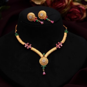May include: A gold-toned necklace and earring set. The necklace features a central circular pendant and is adorned with red, green, and clear gemstones. The matching earrings have a similar design. The jewelry is displayed on a black mannequin head.