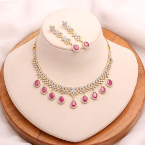May include: A gold-coloured necklace and earring set. The necklace features clear, round-cut stones and teardrop-shaped, ruby-coloured stones. The matching earrings have a similar design. The jewellery is displayed on a mannequin.