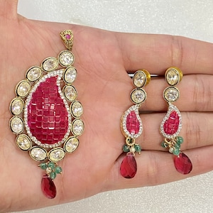 以下が含まれることがあります： クリアな石で囲まれた涙型の赤い石が付いた、ゴールドのペンダントとイヤリングのセット。ペンダントはペイズリー柄で、イヤリングは涙型で、下に緑色の石がぶら下がっています。