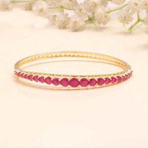 May include: A gold-toned hinged bangle bracelet featuring a row of round, vibrant pinkish-red gemstones set along the front half. The metal band has a polished finish and a structured, open-work design that secures with a simple clasp mechanism.