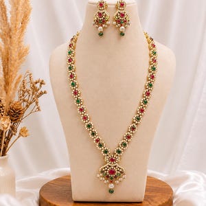 May include: A gold-toned necklace and matching earrings set. The necklace features alternating red and green gemstones, accented with small white pearls. The pendant and earrings have a cluster of gemstones and pearls. The jewelry is displayed on a mannequin.