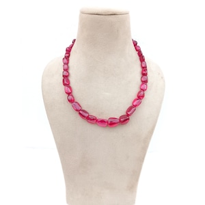 May include: A vibrant necklace featuring polished, oval-shaped ruby gemstones. The beads are strung together, creating a continuous loop. The necklace is displayed on a neutral-toned mannequin bust against a white background, highlighting the rich red color.