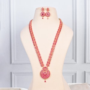Könnte beinhalten: Ein rubinfarbenes Halsketten- und Ohrring-Set, das auf einer Schaufensterpuppe ausgestellt ist. Die Halskette hat ein langes, verziertes Design mit einem großen Anhänger und passenden Ohrringen. Der Schmuck ist in einem goldfarbenen Metall gefasst.