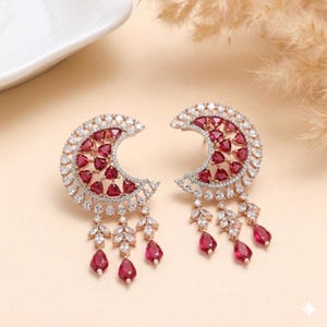 May include: A pair of crescent-shaped earrings with a combination of white and red gemstones. The earrings have a dangling design with teardrop-shaped red gems. The earrings are set against a neutral background.