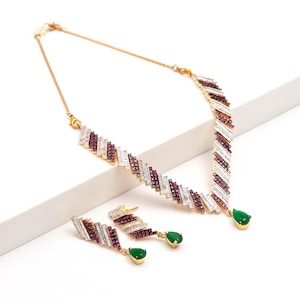 Puede incluir: Un conjunto de collar y pendientes en tono dorado con un patrón de chevrón de piedras claras en corte baguette y pequeñas piedras de color rojo oscuro. El collar y los pendientes terminan en un colgante de piedra verde en forma de pera.