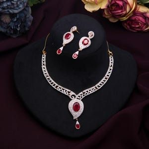 Peut inclure: Un ensemble collier et boucles d'oreilles doré avec une pierre centrale rouge rubis et des pierres blanches. Le collier a un pendentif en forme de goutte et des boucles d'oreilles assorties. L'ensemble est présenté sur un buste noir.