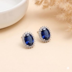 May include: A pair of stud earrings featuring oval, deep blue gemstones surrounded by a halo of smaller, clear stones. The earrings are set in a silver-colored metal and rest on a light beige surface, with a blurred white object and dried plants in the background.