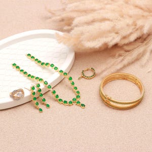 May include: A collection of gold-toned jewelry featuring emerald green gemstones. The set includes a necklace, earrings, a ring, a nose ring, and bangles. The jewelry is arranged on a white tray and a light-colored surface.