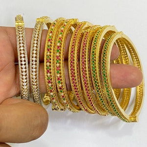 Può includere: Un set di braccialetti d'oro con pietre preziose colorate. I braccialetti sono decorati con file di piccole pietre rotonde in tonalità di verde, rosa e bianco.