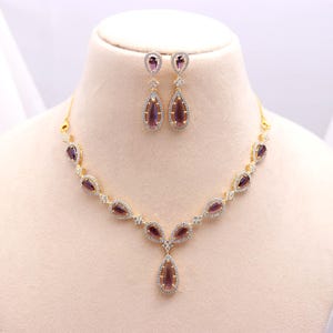 Puede incluir: Un conjunto de collar y pendientes de color dorado. El collar y los pendientes presentan piedras preciosas en forma de lágrima, de color púrpura, cada una rodeada por un halo de pequeñas piedras transparentes. Las joyas se exhiben en un busto de maniquí.