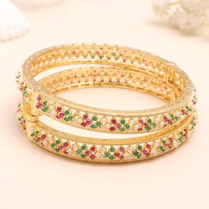 May include: Two gold bangles with intricate detailing. Each bangle is adorned with a pattern of red, green, and white gemstones. The bangles are displayed on a light-colored surface, showcasing their ornate design and craftsmanship.