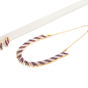Puede incluir: Un collar de tono dorado y un par de pendientes a juego con un patrón diagonal repetitivo de piedras de color rojo oscuro y blanco claro. El collar tiene una cadena fina dorada con cierre de mosquetón y los pendientes son tipo botón.