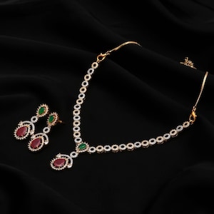 Puede incluir: Un conjunto de collar y pendientes en tono dorado con eslabones circulares incrustados con cristales transparentes. Las piezas de joyería están acentuadas con piedras rojas y verdes en forma de lágrima, con un colgante central en el collar.