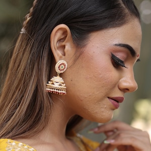 May include: Gold-toned jhumka earrings with a layered bell shape. The earrings feature white pearls, red and green accents, and intricate detailing. The top portion has a circular design with a central green stone. The earrings are a traditional Indian style.