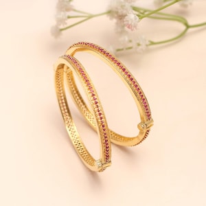 May include: Two gold-coloured bangles, each adorned with a line of small, red gemstones. The bangles are displayed against a soft, neutral backdrop with delicate white flowers. The jewellery has a classic design.