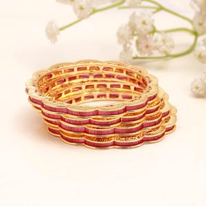 May include: A stack of four gold-toned bangles featuring a scalloped edge design. Each bangle is inlaid with a continuous row of small, rectangular, deep pink stones that wrap around the entire circumference of the circular metal jewellery pieces.