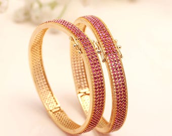Könnte beinhalten: Zwei goldene Armreifen mit strukturiertem Design und einer Reihe kleiner, rosa Edelsteine. Die Armreifen sind miteinander verbunden und vor einem weichen, neutralen Hintergrund platziert. Der Schmuck ist elegant und auffällig.