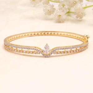 May include: A gold-toned hinged bangle bracelet featuring a central pear-shaped clear stone flanked by two rows of small, pavé-set clear stones. The metal band has a polished finish and a secure clasp mechanism on the side for easy opening.