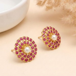 May include: A pair of gold-colored stud earrings. Each earring features a central white pearl surrounded by a ring of small, round, red gemstones. The earrings have a floral design and are set against a light beige background.