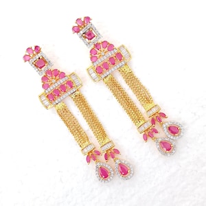 May include: A pair of gold-coloured earrings with pink and clear gemstones. The earrings have a rectangular top with a square pink stone and a floral design of pink stones. Below are gold chains and teardrop-shaped pink stones.