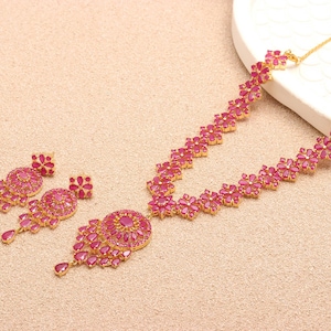 Op de afbeelding: Een goudkleurige sieradenset met een ketting en bijpassende oorbellen. De ketting en oorbellen zijn versierd met helder roze edelstenen, gerangschikt in bloem- en druppelvormige patronen. De set wordt gepresenteerd op een neutrale ondergrond.