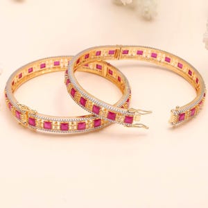 May include: Two gold-toned hinged bangles featuring a series of square-cut vibrant pink stones alternating with small clear accent stones. Each bracelet has a secure metal clasp mechanism and a polished finish, showcasing a repeating geometric pattern.