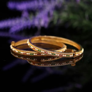 Puede incluir: Dos brazaletes dorados con detalles intrincados. Cada brazalete está adornado con pequeñas cuentas de colores rojo, verde y blanco, reflejadas en una superficie oscura. Los brazaletes son una pieza de joyería clásica.