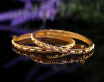May include: A pair of gold-toned hinged bangles featuring a textured surface inlaid with small white pearls, red stones, and green stones arranged in a repeating pattern along the outer circumference of each circular bracelet.