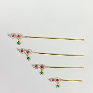 Puede incluir: Cuatro palos de pelo dorados con extremos decorativos. Cada extremo presenta un diseño de dos círculos con una gema rosa en el centro, rodeada de gemas transparentes más pequeñas. Una sola gema verde cuelga de la parte inferior de cada diseño.