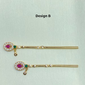May include: Two gold-colored, decorative sticks with a round, jeweled design at the top. Each stick has a small, green stone hanging from the top design. The text "Design B" is visible at the top of the image.