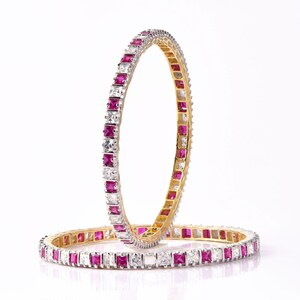 Könnte beinhalten: Zwei goldene Armreifen mit einer Reihe von quadratischen rosa und weißen Steinen.