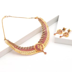 Puede incluir: Un conjunto de collar y pendientes colgantes en tono dorado con intrincados trabajos de filigrana metálica y pequeñas piedras rosas incrustadas. El collar tiene un diseño curvo en forma de media luna con un colgante circular central.