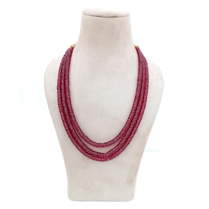 May include: A three-strand necklace with a deep red hue, displayed on a mannequin. The necklace appears to be made of small, round beads, creating a textured surface. The strands are layered, adding depth to the design.