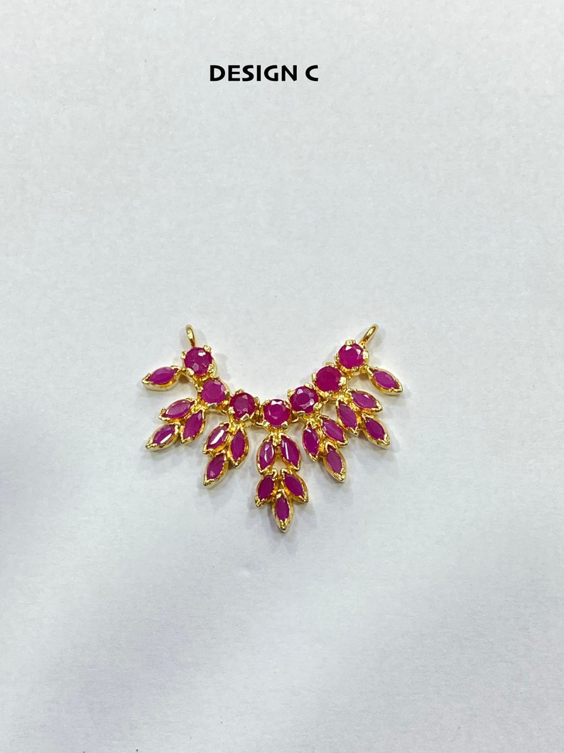 May include: A gold-toned jewelry connector labeled DESIGN C, featuring a symmetrical arrangement of vibrant fuchsia-pink marquise and round-cut stones set in a leaf-like pattern with two small loops at the top for attachment to chains or cords.