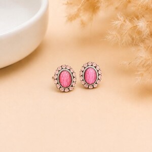 May include: A pair of oval stud earrings featuring a central carved pink stone with a floral motif, surrounded by a halo of smaller, lighter pink faceted stones set in a metallic frame with a dark, oxidised finish around the centre stone.