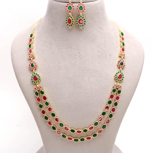 May include: A gold-coloured necklace and earring set. The necklace has two strands of alternating red and green oval-shaped stones. The earrings and necklace have a floral design with red and green stones. The jewellery is displayed on a mannequin.
